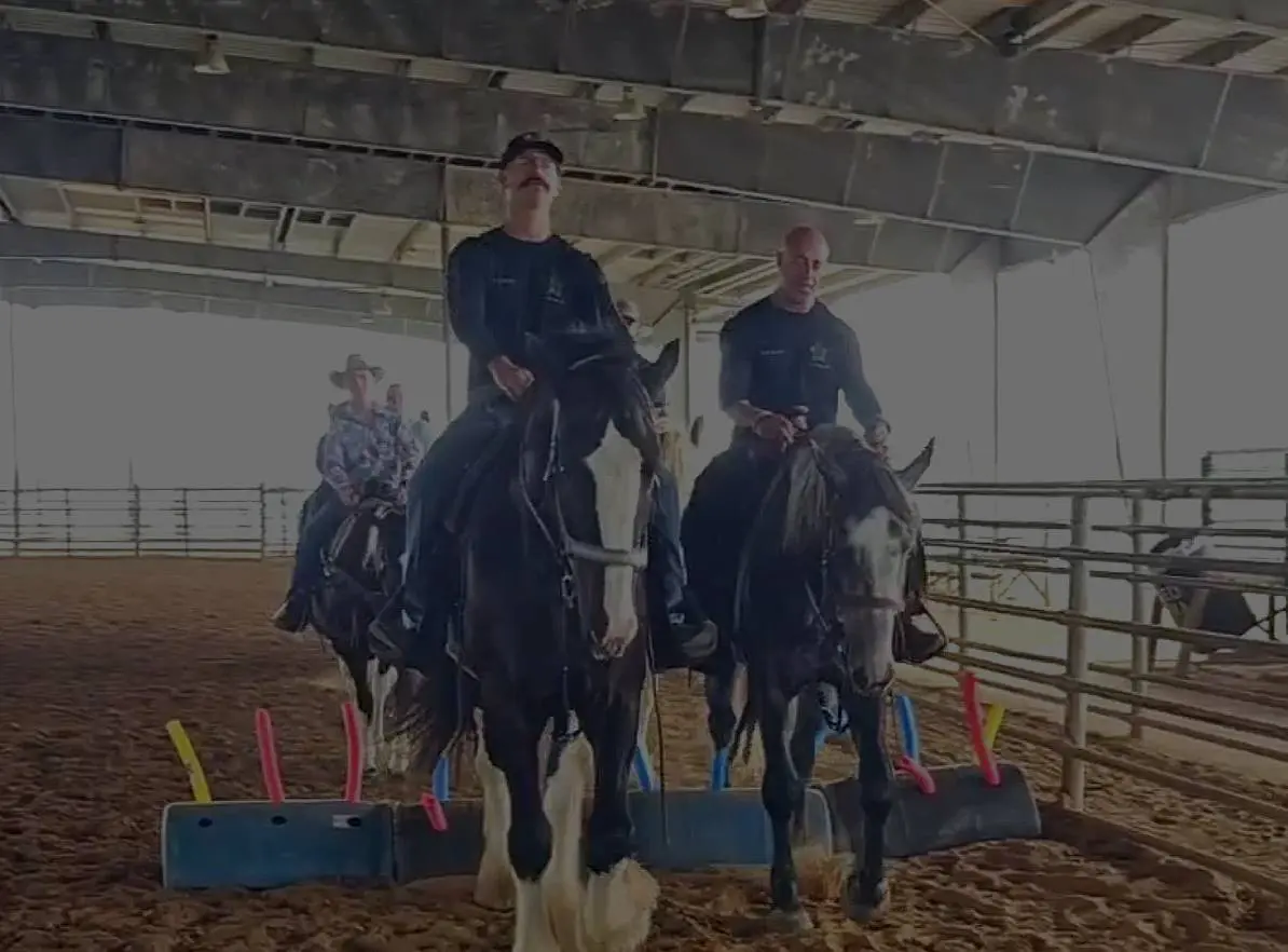 Two people riding horses indoors in a fenced arena.