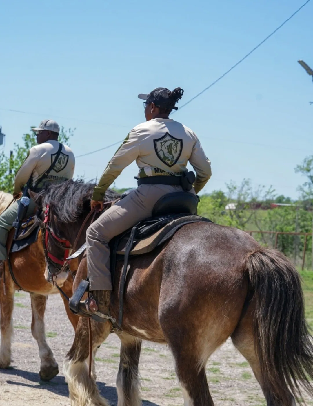 Two riders on horseback in a rural setting under a clear sky.